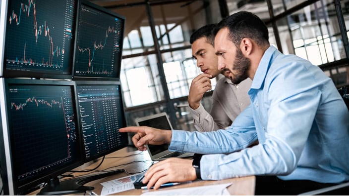 Two male office workers looking at four large computer monitors.