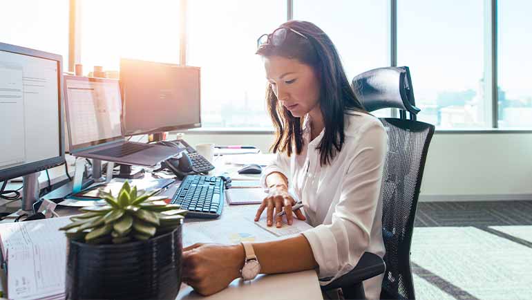 Woman at her desk in an office with 3 computers on her desk looking at treasury exchange rates