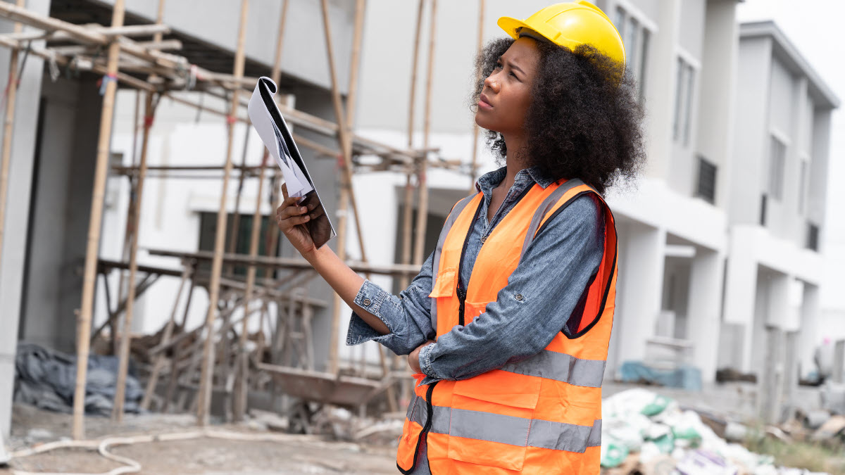 Photo of a woman wearing a hardhat and vest and holding a clipboard as she observes a construction site.