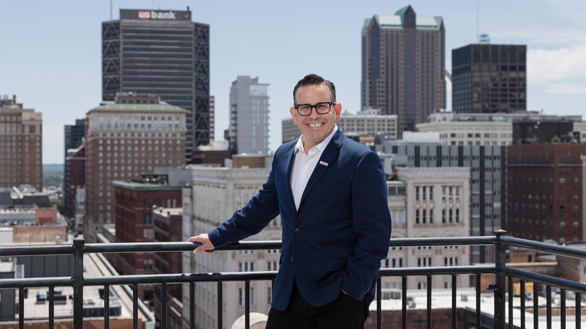 Man in suit standing on rooftop with skyline behind him.