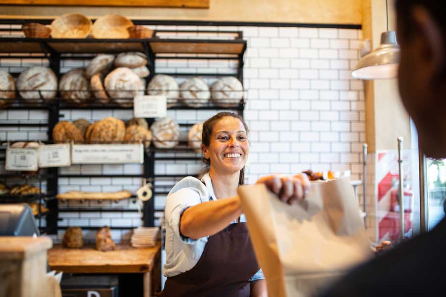 Owner of a bakery handing a bag of purchased goods to a customer.