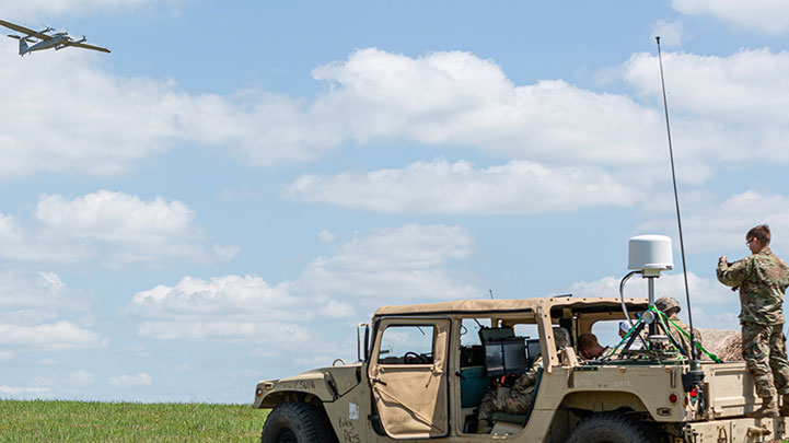 Army soldiers sitting in a Humvee in an open field while a military plane flies overhead.