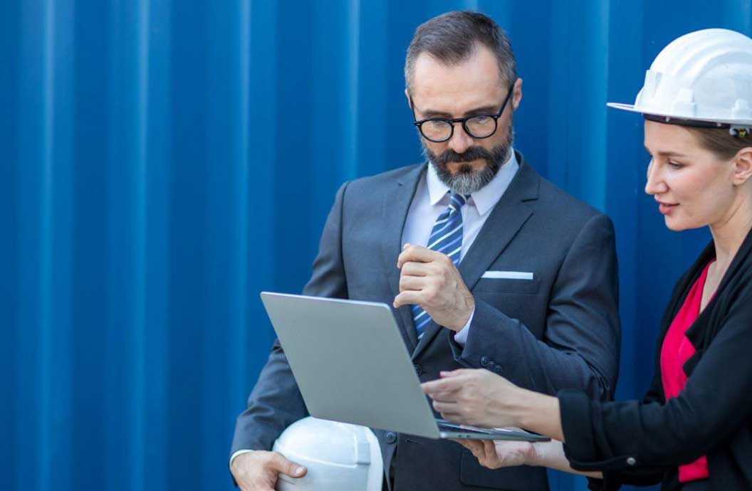 Woman wearing a hardhat standing next to man in a suit. She is pointing to a laptop as they discuss something.