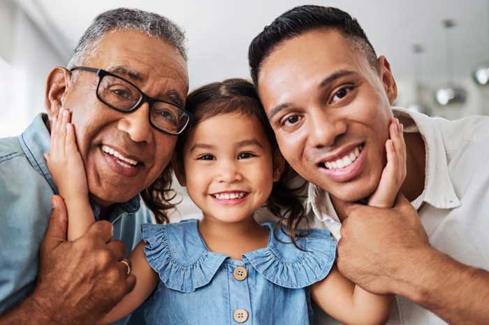 Tres generaciones de una familia hispana sonríen para la cámara: una niña en el medio, su abuelo a la izquierda y su padre a la derecha.