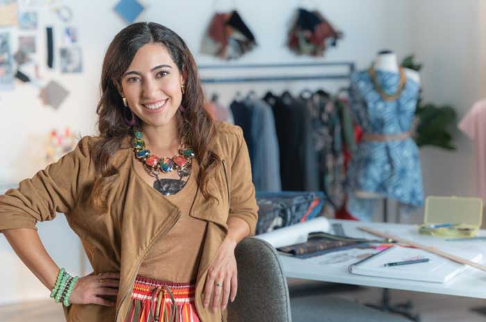 Un joven diseñador de ropa hispano posa delante de su lugar de trabajo. En el fondo se ve ropa en perchas y un vestido en proceso de confección.