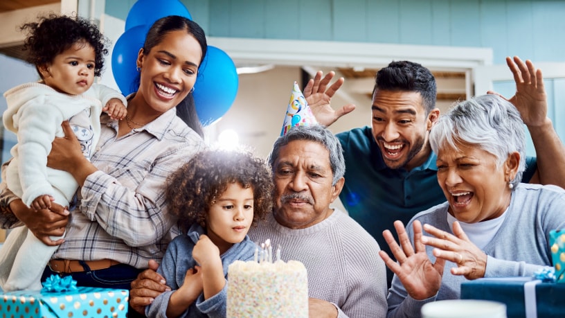 Una madre con dos niños(as) celebrando el cumpleaños de papá con un pastel y gorros para fiesta.