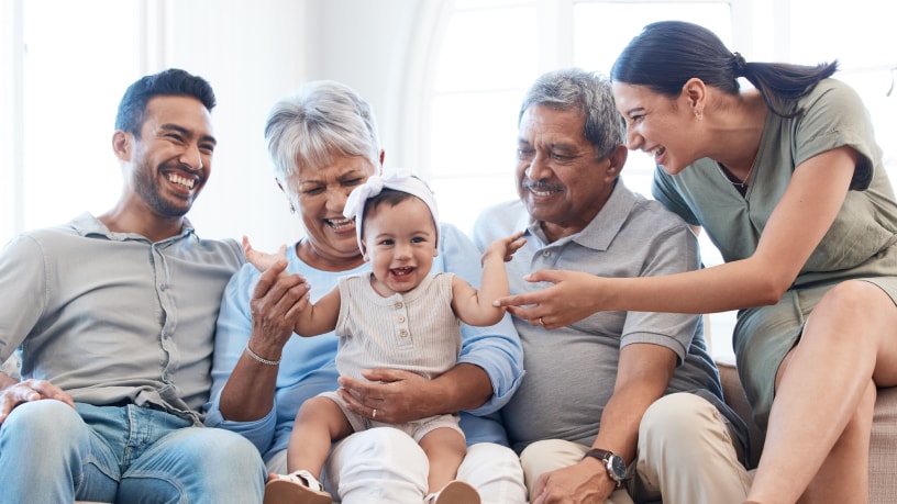 Tres generaciones de una familia hispana sonríen y se ríen juntas, y todos los adultos se centran en la pequeña en el regazo de su abuela.